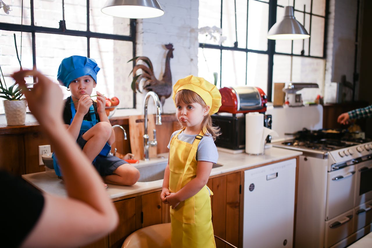 gallery-03 Two kids in colorful chef hats enjoy a playful cooking session in a cozy kitchen setting.