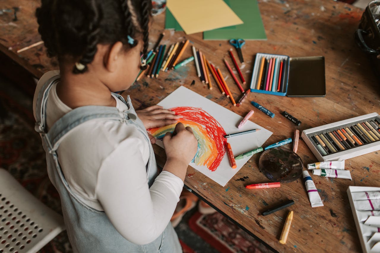 services-03 Young child creating a vibrant rainbow drawing at a wooden table with various art supplies.